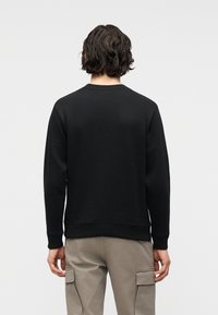 Νέος/νέα με κυματιστά σκούρα μαλλιά, που φοράει μαύρο φούτερ με μακριά μανίκια και ανοιχτό γκρι παντελόνι cargo, στέκεται γυρισμένος/η πλάτη.