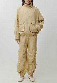 Beige Nylon-Trainingsanzug mit einer Zip-Jacke, die zwei Fronttaschen und elastische Bündchen hat, kombiniert mit lockeren Hosen und elastischen Knöchelbündchen.