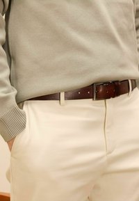 Bruine leren riem met zilveren gesp, gedragen over lichtgekleurde broek en een gestructureerde lichtgrijze trui met geribde boorden.