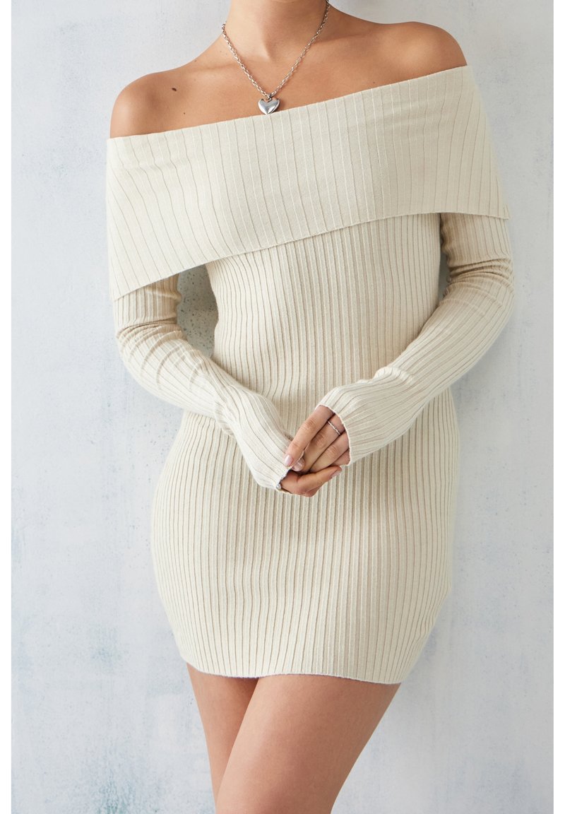 Femme portant une robe en maille côtelée beige à épaules dénudées avec manches longues et un collier pendentif cœur en argent, les mains jointes devant elle.