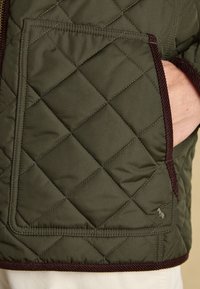 Hand in Tasche einer olivgrünen gesteppte Jacke mit braunem Besatz und gesticktem kleinen Tierlogo nahe der unteren rechten Ecke.