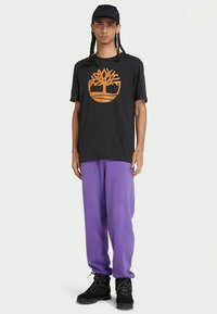 Sort bomuld T-shirt med et orange trælogo, parret med løse lilla sweats og sorte støvler. Simpelt design, afslappet pasform.
