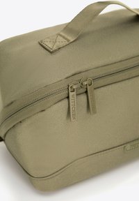 Oliwkowa kosmetyczka z fakturowaną powierzchnią, uchwytem u góry oraz podwójnymi suwakami z logo marki. Lekko zwężający się kształt.