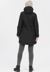 Svart vattentät jacka med huva, med insvängd midja, långa ärmar och medellång design. Bärs över grå byxor och stövlar.