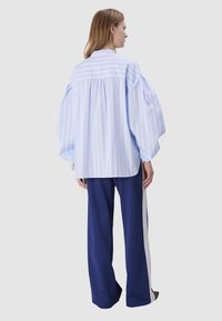 Chemise oversize rayée bleu clair avec épaules arrondies et dos froncé, associée à un pantalon large bleu marine avec une rayure blanche sur le côté.