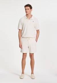 Beige, rövidujjú pólóing kék logóval, melyet hasonló bézs színű, zsinóros rövidnadrággal viselnek, könnyű, slip-on cipőkkel.