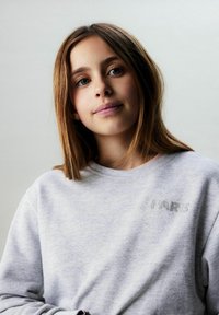 Gray sweatshirt with the word "PARIS" embroidered in shiny material, worn by a person with brown hair and blue eyes. Soft texture, round neckline.