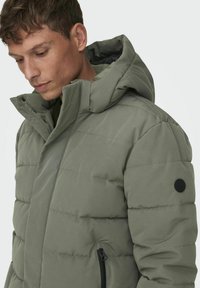 Pufferjack in olijfgroen met een gewatteerd patroon, hoge kraag en zwarte accentknop. Voorzien van ritszakken en een ruime capuchon.