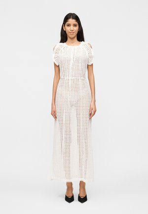 Vrouw die een wit, opengeweven maxi-jurk draagt met pofmouwen en uitsparingen bij de schouders, gecombineerd met zwarte schoenen met een spits uiteinde.