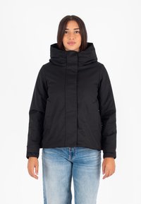 Vrouw in een zwarte hoodie met hoge kraag en lichtblauwe jeans, staand tegen een witte achtergrond.
