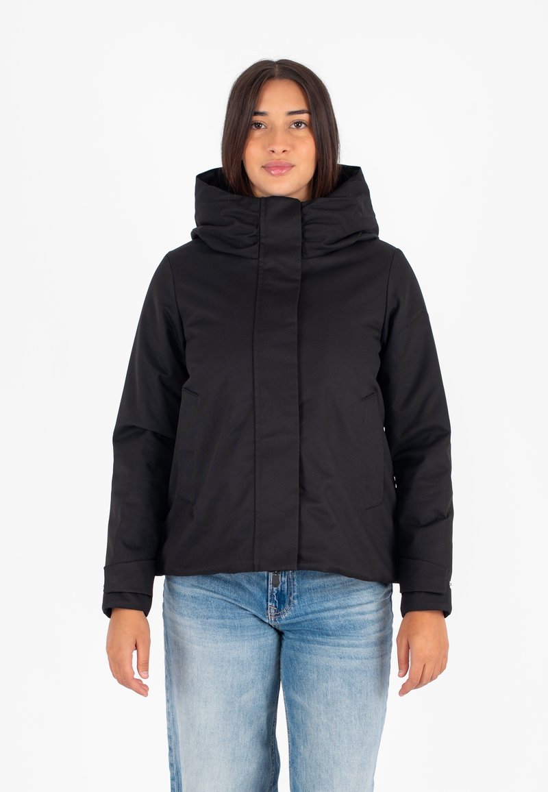 Vrouw in een zwarte hoodie met hoge kraag en lichtblauwe jeans, staand tegen een witte achtergrond.