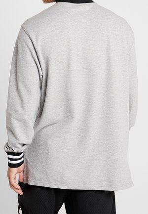 Mężczyzna ubrany w jasnoszary sweter z długim rękawem z czarno-białymi paskowanymi mankietami, pokazany od tyłu na jednolitym tle.