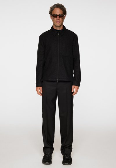 Svart ullblandad zip-up jacka med krage och en bröstficka. I kombination med svarta byxor och svarta sneakers. Man som bär solglasögon.