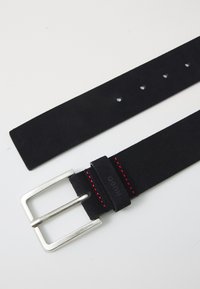 Curea din suede negru cu o cataramă din metal argintiu. Prezintă cusături roșii și mai multe orificii pentru ajustare. Textură netedă pe toată lungimea.