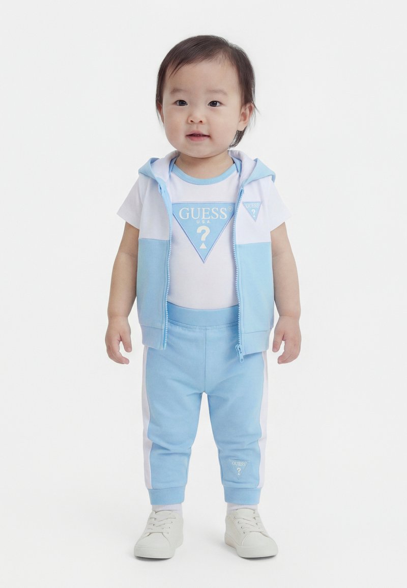 Νήπιο που φοράει λευκή και γαλάζια στολή Guess με γιλέκο με κουκούλα, παντελόνια και λευκά αθλητικά παπούτσια, στέκεται μπροστά σε λευκό φόντο.