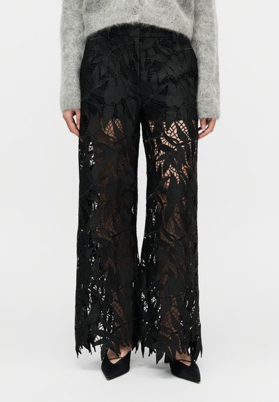 Pantalon en dentelle noire avec motif de feuilles, coupe large, texture transparente et bords bruts, associé à des escarpins noirs pointus.