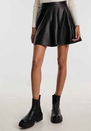 Kobieta ubrana w czarną skórzaną mini spódniczkę, beżowy prążkowany sweter oraz czarne masywne botki, stojąca na jasnym tle.