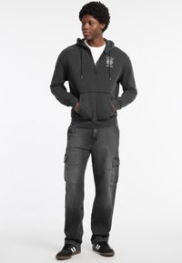 Felpa zip grigia scura con tasche frontali, abbinata a pantaloni cargo grigi scuri con tasche laterali. Sneakers nere per un contrasto.