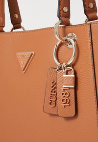 Borsa a tracolla beige con finitura testurizzata, dotata di hardware dorato, un logo triangolare e etichette portachiavi impresse con "GUESS" e "1981."
