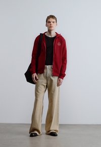 Červená mikina na zips s logom, čierne tričko, béžové široké nohavice, čierne topánky a čierna taška. Vnútorné prostredie s jednoduchým pozadím.