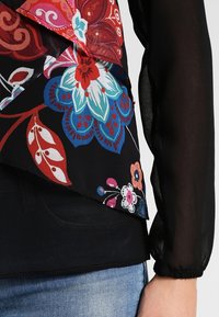 Blouse florale multicolore avec un voile noir transparent, présentant des motifs vifs et un design en couches ; manches longues bouffantes avec poignets élastiques.