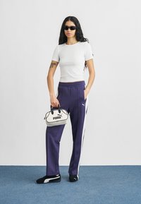 Μωβ φόρμες με λευκές πλευρικές ρίγες, εφαρμοστό λευκό μπλουζάκι, μαύρα αθλητικά παπούτσια και μια μικρή κρεμ τσάντα χειρός με μαύρο λογότυπο.