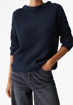 Persona vistiendo un suéter de punto grueso azul marino con las mangas remangadas y jeans gris oscuro, con una mano en el bolsillo sobre un fondo blanco.