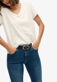 Camiseta blanca de cuello en v hecha de una tela suave, combinada con jeans de mezclilla azules y un cinturón negro con un detalle de hebilla dorada. Manos en los bolsillos.