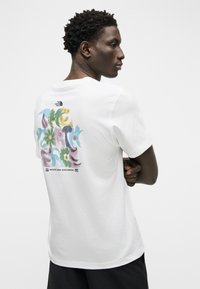 Άνδρας με σταυρωμένα χέρια, φορώντας λευκό μπλουζάκι The North Face με πολύχρωμα αφηρημένα ανθικά γραφικά και το κείμενο "Never Stop Exploring" στην πλάτη.