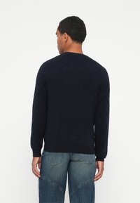 Hombre de espaldas, vistiendo un suéter texturizado en azul marino oscuro y jeans azules desgastados con detalles de costura diagonal en la parte trasera de las piernas.