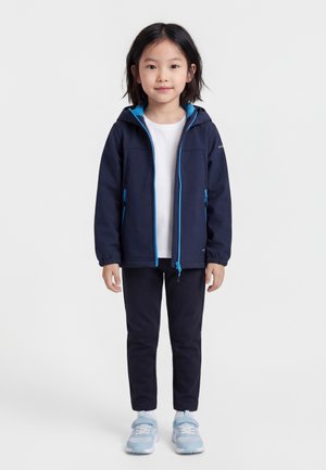 Bambino in piedi, che indossa una giacca con cappuccio blu navy, maglietta bianca, pantaloni blu navy e scarpe da ginnastica azzurri su uno sfondo neutro.