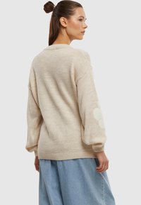 Beiger Strickpullover mit lässiger Passform, der Ballonärmel und sanfte Blumenmuster an den Ellenbogen aufweist. Über einem hellblauen Rock getragen.