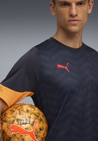 Niebieska i pomarańczowa koszulka sportowa z wzorem w zygzak i logo Puma. Pomarańczowa piłka do futbolu z geometrycznym wzorem i pasującym logo.