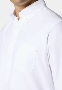 Bílá košile s dlouhým rukávem, vybavená límcem na knoflíky, kapsou na levé hrudi a texturovanou tkaninou s jemným leskem.