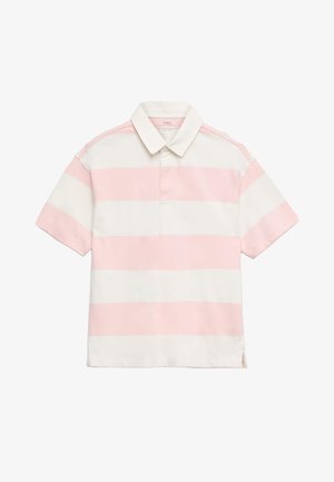 Młoda osoba stoi ze skrzyżowanymi ramionami, ubrana w różowo-białą koszulkę polo w paski, niebieskie szorty i białe trampki na jednolitym tle.