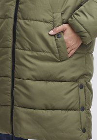 Olivově zelená prošívaná bunda s zipovým zapínáním, bočními kapsami a černými patentkami na lemu. Hladký povrch s prošívanými panely.