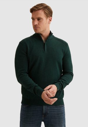Hombre con pelo castaño claro corto que lleva un suéter verde oscuro con cremallera hasta el cuarto y vaqueros azules, está de pie con las manos entrelazadas.