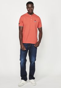 Korálové polo tričko s henley límcem a třemi knoflíky, kombinované s tmavě modrými džínami a bílými teniskami. Na hrudi je umístěno logo.