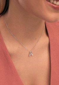 Collar de plata con un colgante en forma de letra "A" adornado con un diamante, exhibido sobre un cuello desnudo contra un fondo rosa.
