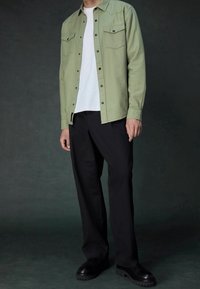 Camicia button-up verde chiaro con taschini sul petto, indossata sopra una maglietta bianca, abbinata a pantaloni neri e scarpe nere. Texture liscia.