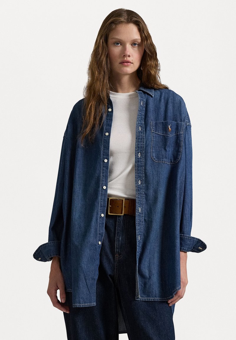 Camicia di denim di colore blu scuro, vestibilità comoda con maniche lunghe, apertura con bottoni davanti e taschino frontale singolo. Indossata sopra una maglietta bianca.