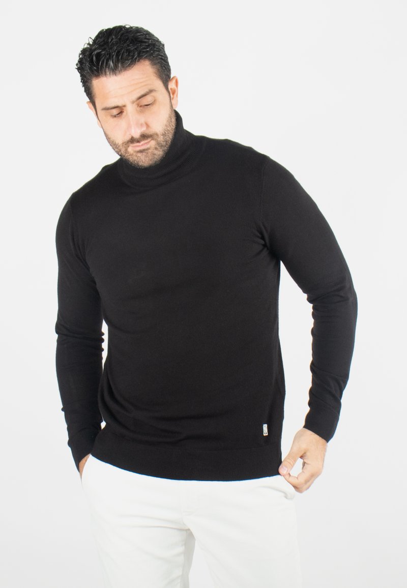 Pull à col roulé noir avec manches longues, poignets et ourlet côtelés. Texture lisse, coupe ajustée, associé à un pantalon blanc.