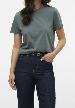 Vrouw draagt een gedempt groene korte mouw T-shirt in een hooggetailleerde donkerblauwe jeans met een zwarte riem, hand in zak.
