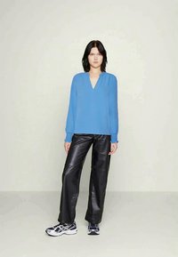 Mujer vestida con una blusa azul de manga larga, pantalones de cuero negros y zapatillas en blanco y negro, de pie contra un fondo claro y liso.