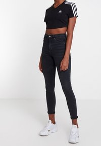 Černé zkrácené tričko s bílým designem tří pruhů a logem. Černé úzké džíny s vysokým pasem. Bílé tenisky se zlatými akcenty.