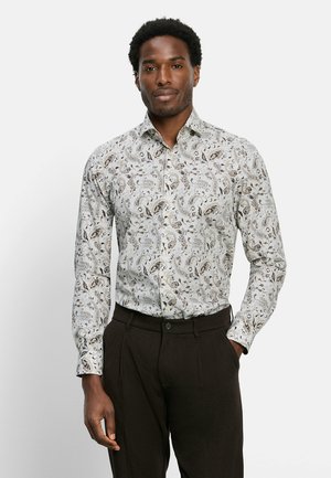 Mann steht, trägt ein weißes Langarmhemd mit braunen Paisley-Mustern und dunkelbraune Hose vor einem einfarbigen hellen Hintergrund.