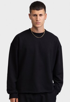 Νέος άνδρας με κοντά σκούρα μαλλιά φοράει μαύρο φούτερ με στρογγυλή λαιμόκοψη, χρυσή αλυσίδα στο λαιμό και μικρούς κρίκους στα αυτιά, στέκεται μπροστά σε φωτεινό φόντο.