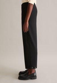 Schwarze, maßgeschneiderte Hose im geraden Schnitt mit glatter Textur, seitlichen Taschen und Knopfakzenten am Saum. Kombiniert mit schwarzen Slippern.