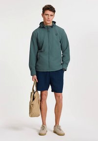 Jachetă ușoară pentru bărbați, de culoare teal, cu fermoar în față, glugă ajustată și logo. Asortată cu pantaloni scurți bleumarin cu dungi și adidași bej, ținând o geantă maro deschis.