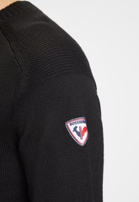 Schwarzer Strickpullover mit strukturierter Stoffoberfläche. Mit einem bunten Rossignol-Logo-Patch am linken Ärmel. Rundhalsausschnitt und gerippte Bündchen.